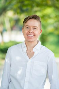 A white person with short, blonde hair and blue eyes is wearing a white button-down shirt in the foreground. There are blurred trees in the background.